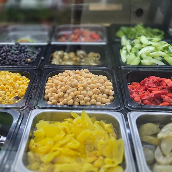 A deli salad bar with various toppings including chickpeas, corn, black beans, and roasted red peppers.