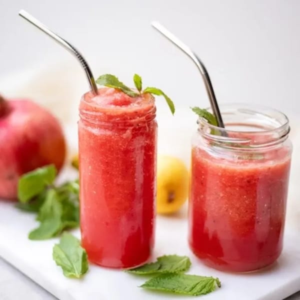 Two red fruit smoothies in glass jars with metal straws, garnished with mint, on a white surface.
